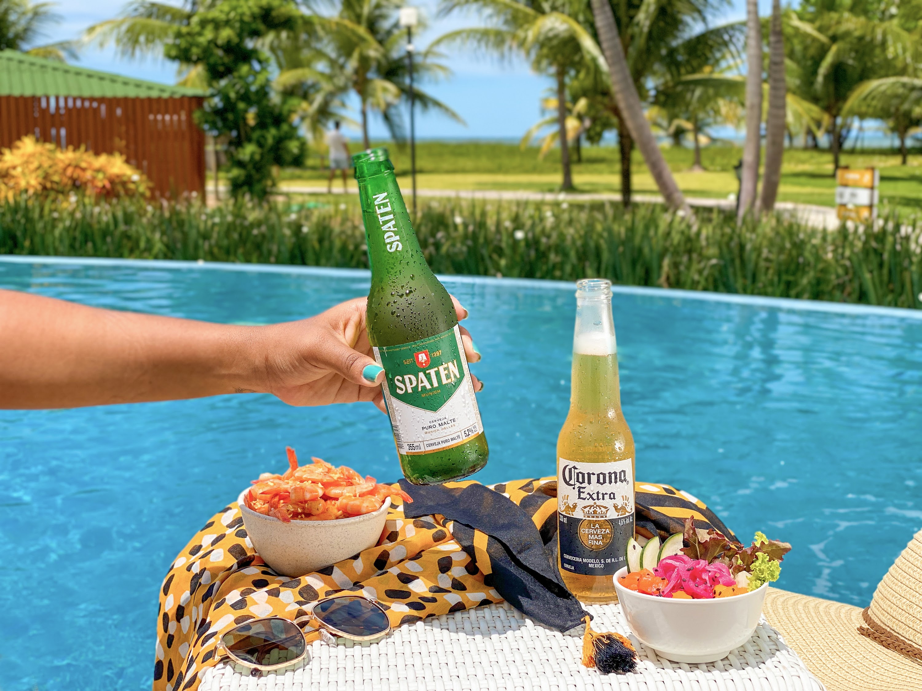 Pratos de petiscos a lado de duas cervejas long neck, uma Spaten e uma Corona Extra, em mesa na piscina do Salinas Maragogi